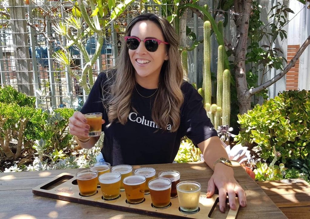Image of Nadia having a great Perth Weekend. She in sunglasses and a black shirt. She is holding a beer and has a tasting paddle in front of her. Behind her are trees and plants.