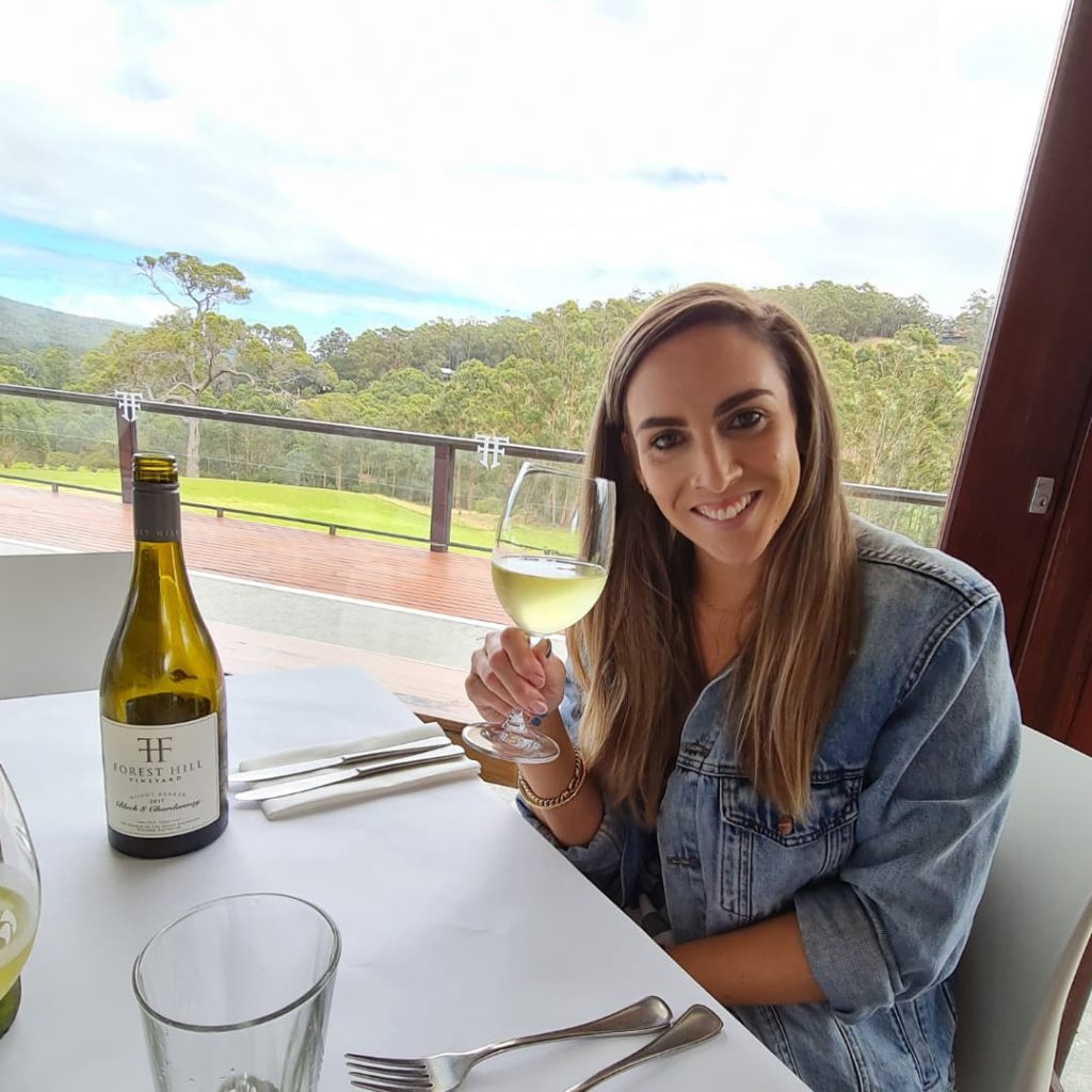 Nadia sitting at a table holding a glass of white wine. She is smiling at the camera. Behind her is a field of grass leading to trees. 