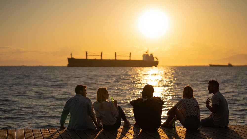5 friends drinking beer and sitting at the edge of the water at sunset. There is a ship in the distance out at sea