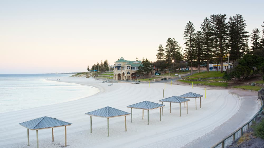 Image of Cottesloe beach in early morning. A great Perth Weekend spot. Beach shelters are on the sand, and the beach is framed by large pine trees on the right