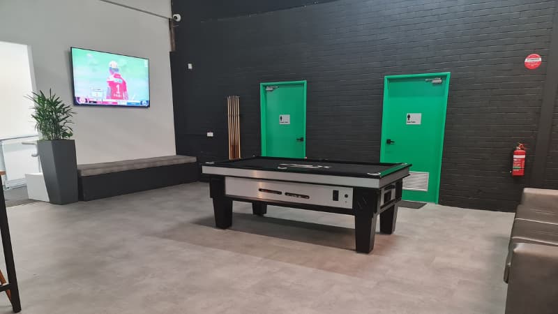 Image of pool table and TV with the cricket on. There are bathrooms in the background.