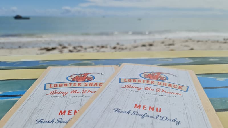 Two lobster shack menus sitting on a surfboard table with the beach in the background