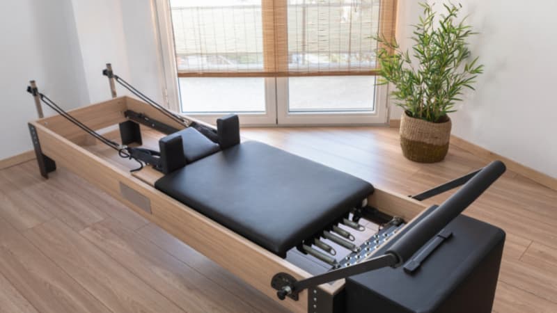 Image of a pilates reformer on a wood floor. There is a window in the background and a plant to the right