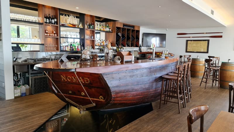Image of the bar that is a converted yacht at Colourpatch cafe. There are wood bar stools surrounding it, barrel tables and shelves with sprits at the back