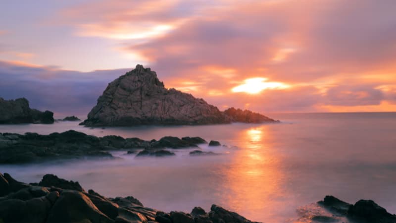 The sun is setting over Sugarloaf Rock creating a pink and purple sky which is reflected in the ocean.