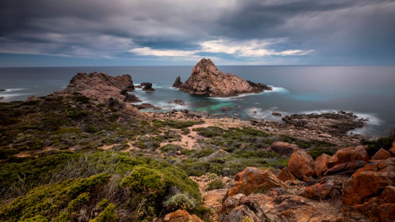 Sugarloaf rock on an overcast day. Clouds int he sky are dark grey and are casting shadows on the green shrubs in the foreground.