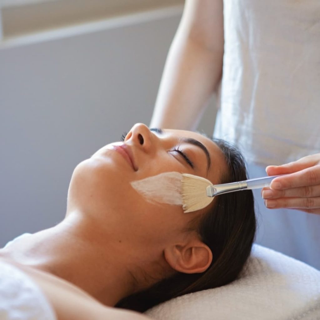 Woman laying on treatment bed having a facial treatment applied to her face with a brush