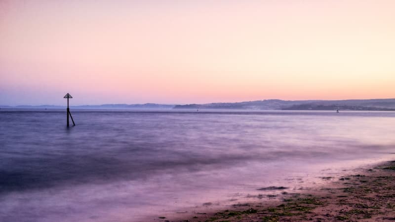 A sign pokes out of the water of an Exmouth beach at sunset. The ocean is reflecting the purple colours of the sky.