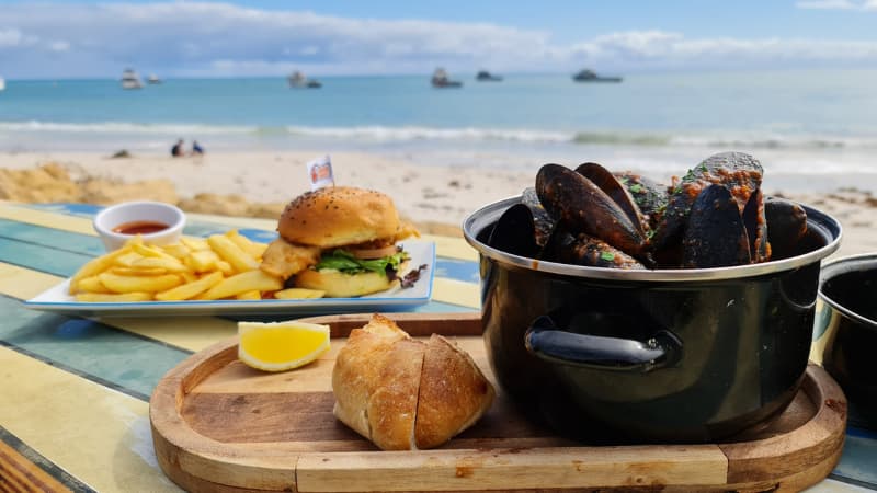 A pot of chilli mussels and a fish burger sit on a table in front of the ocean at Lobster Shack