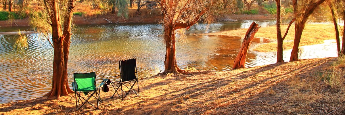 Two camp chairs sit along the riverbank as golden sunlight shines through the trees.