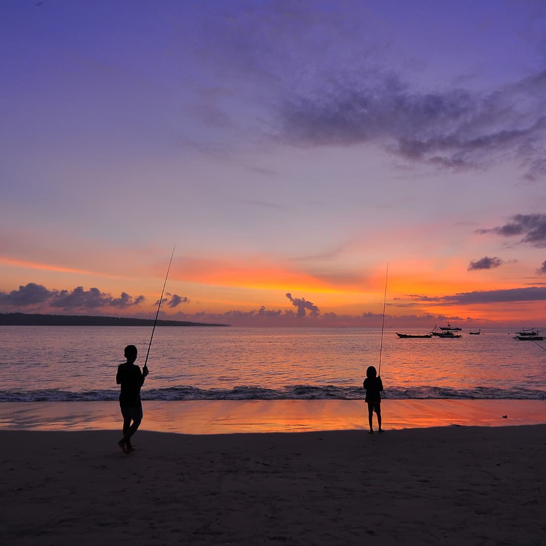 Two people stand on a beach at sunset fishing. The sky is orange and purple.