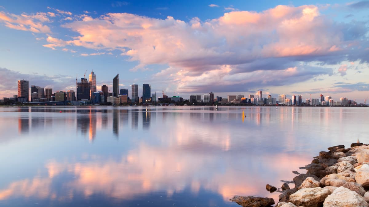 The Perth city skyline across from the Swan River at sunrise. The sky and reflection in the water are soft pinks and blues.