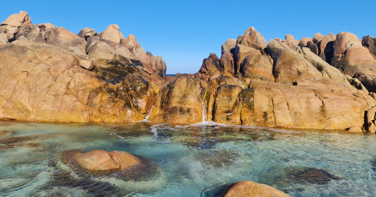 A view of Injidup Natural Spa, showcasing smooth, golden-brown rock formations rising from crystal-clear turquoise water. Small streams of water cascade down the rocks, creating a tranquil scene under a bright blue sky.