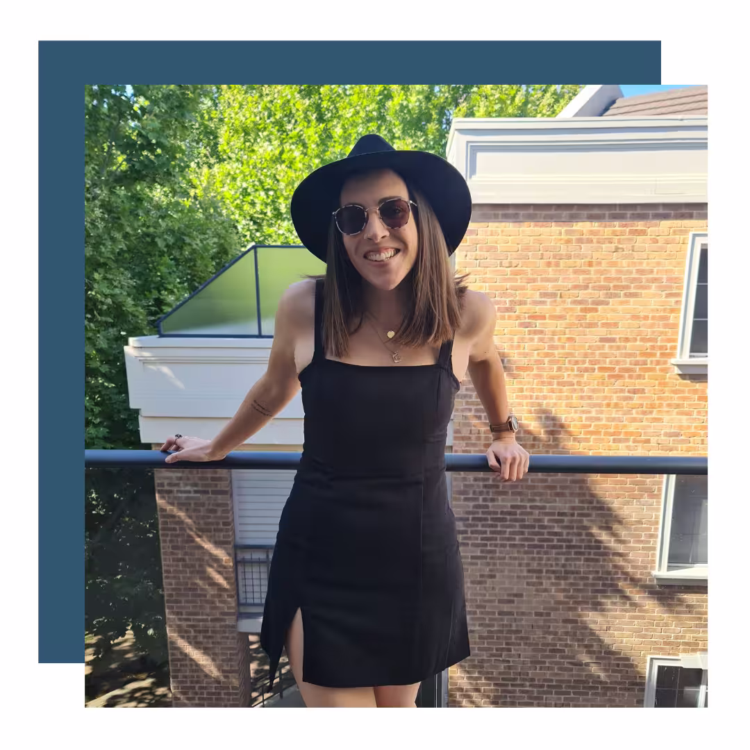 Nadia Perth Weekend Nadia standing on a balcony in a black dress and wide-brimmed hat, smiling at the camera with sunglasses on. Background features a sunny day with green trees and a brick building.