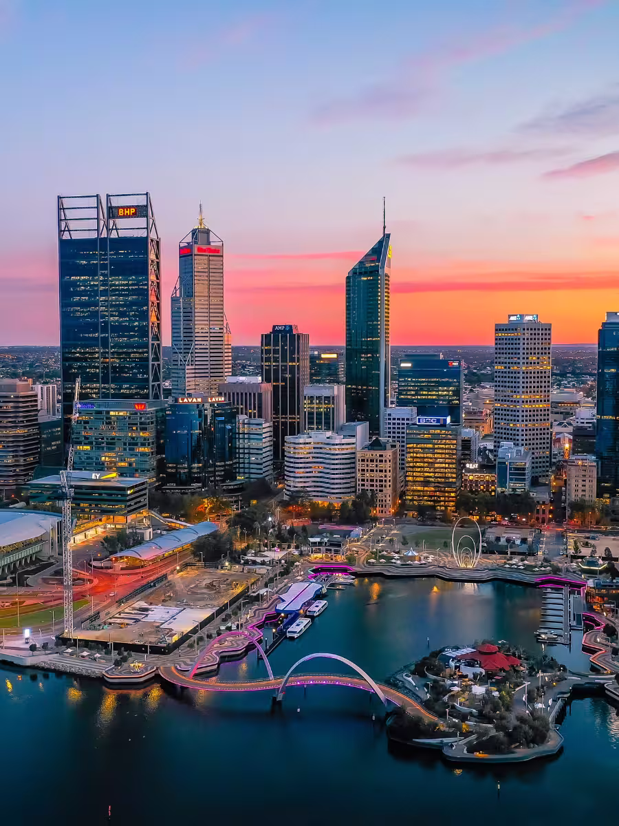 Perth Sunset Skyline Drone view of Elizabeth Quay and the Perth city skyline at dusk, with colourful lights reflecting off the water and the illuminated pedestrian bridge.