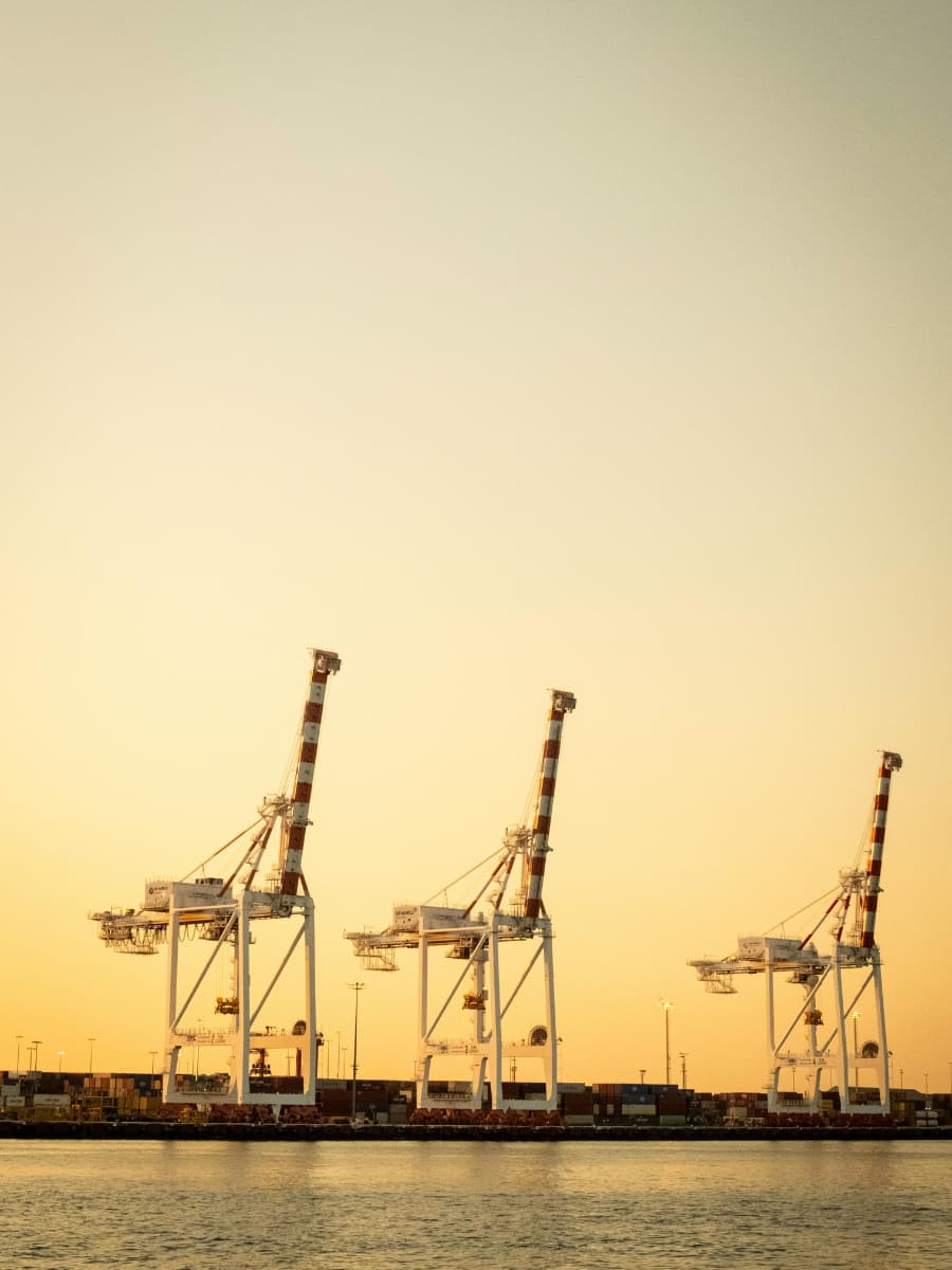 Fremantle Port Three large container cranes at Fremantle Port silhouetted against a soft orange sunset sky.