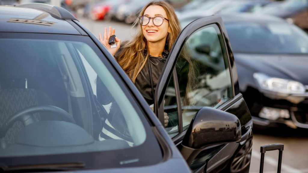 Smiling woman holding car keys while getting into a rental car at an airport car park